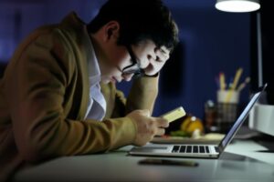 Hombre cansado usando teléfono y laptop, ilustrando tecnoadicción