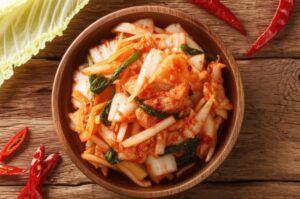 Kimchi casero en un tazón de madera sobre una mesa de madera, con hojas de col y pimientos rojos.