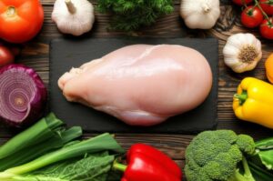 Pechuga de pollo sin piel sobre tabla de cortar negra, rodeada de verduras frescas.