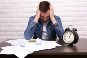 Hombre frustrado sentado en un escritorio con papeles arrugados y un reloj, representando la procrastinación