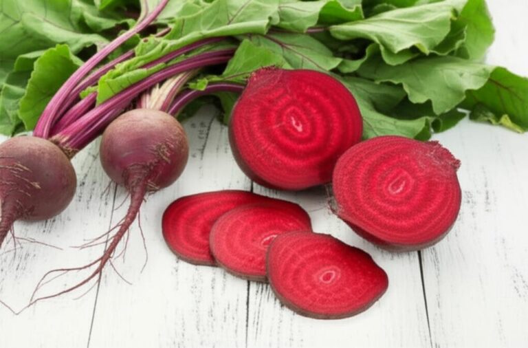 Remolachas frescas enteras con hojas y cortadas, exhibiendo su vibrante color rojo sobre superficie de madera clara.