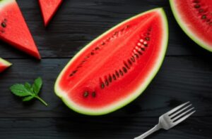 Sandía cortada sobre mesa oscura con tenedor y hojas de menta, lista para consumir.